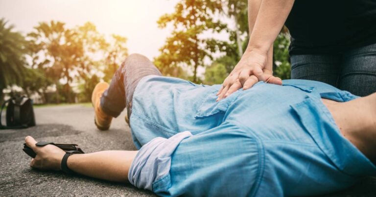 A bystander giving an unconscious male CPR in the middle of a street is pressing into the male’s chest with their hand