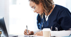 A female healthcare professional writing in a notebook. She has a laptop, stack of books, and stethoscope near her.