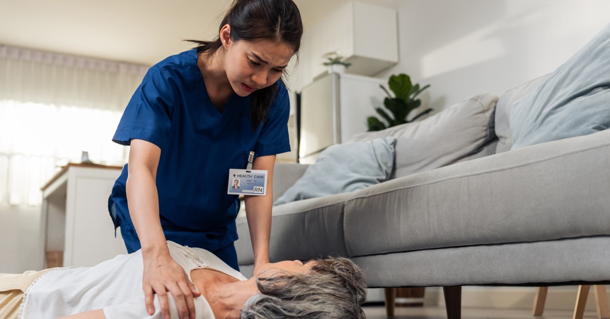 A caregiver kneeling beside and gently shaking the shoulders of an older adult patient inside of the patient's home.