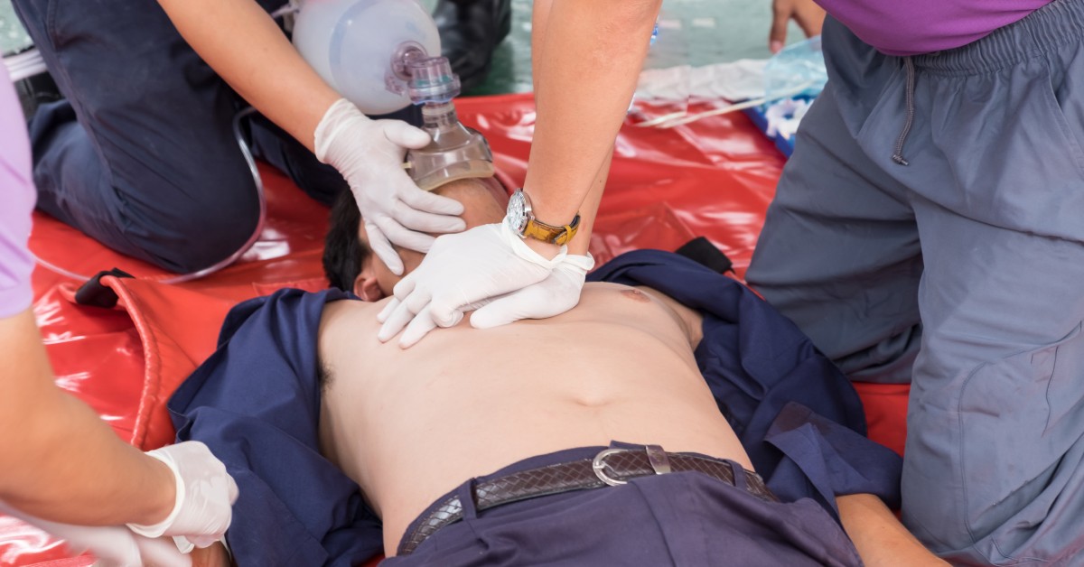 A team of three medical professionals and an adult male laying on an orange tarp. The team performs CPR on the man.