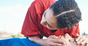 On a playing field, a woman listens for an unconscious athlete's breathing and prepares to perform CPR.