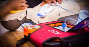 A person is using a practice mannequin for AED training. To the right is a red training pack with text reading, "AED."