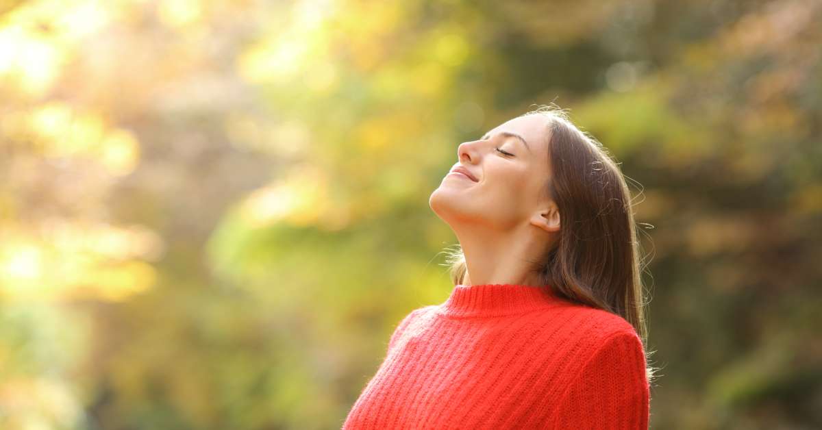 A smiling woman in a red turtleneck sweater has her eyes closed while taking a deep breath in the woods on a sunny day.