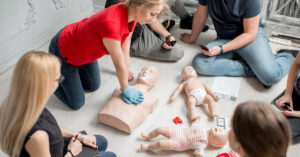 A young woman uses a CPR dummy to demonstrate chest compressions to a group of students sitting on the ground.