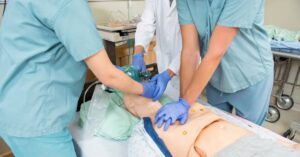 Two people wearing blue scrubs and one wearing a white coat practice CPR on a dummy in a hospital room.