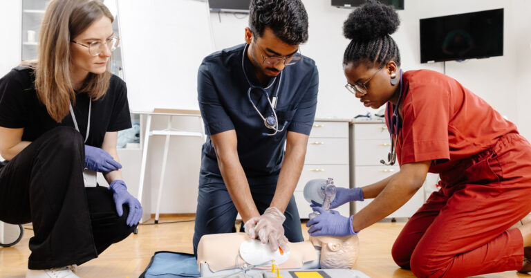 Three people wearing different colored scrubs kneel on a wooden floor as one practices CPR techniques on a dummy.