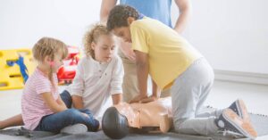 Three young children practice CPR on a dummy while an instructor in a light blue shirt observes them in a classroom.