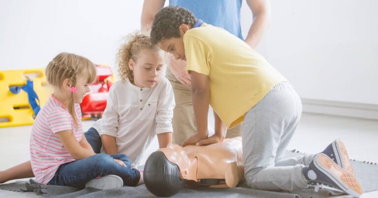 Three young children practice CPR on a dummy while an instructor in a light blue shirt observes them in a classroom.