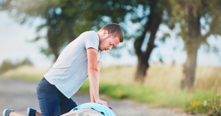 A man wearing a white tee shirt kneeling on the ground while performing chest compressions on someone lying down.