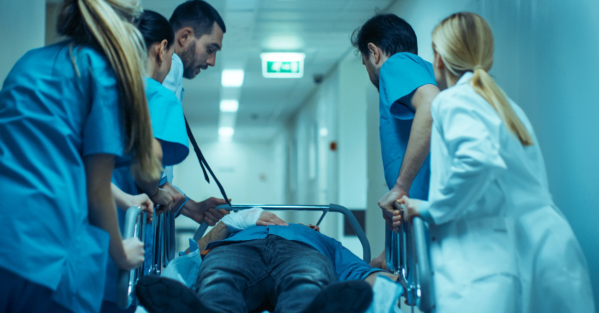 Four people wearing blue scrubs and one wearing a white doctor's coat wheel a patient on a bed down a hospital hallway.