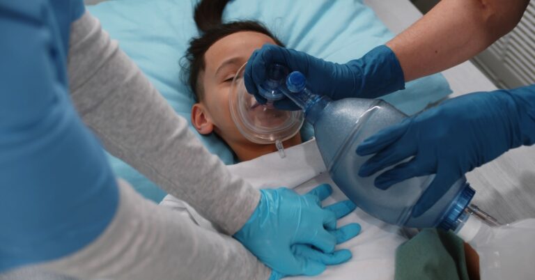 A close-up showing a pair of medical professionals wearing blue scrubs and blue gloves using a tube to perform CPR on a child.