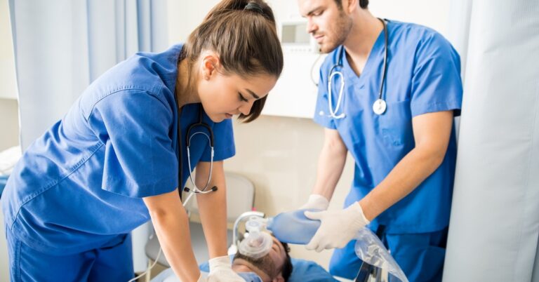 A man and a woman wearing blue scrubs and white plastic gloves perform CPR on an unconscious man laying down.