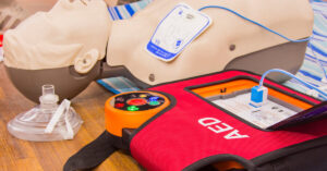 A close-up of an automated external defibrillator (AED) with its pads attached to a practice dummy on a wooden floor.