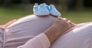 A close-up of a pregnant belly in pink maternity wear with blue-baby shoes on top and hands cradling the bump.