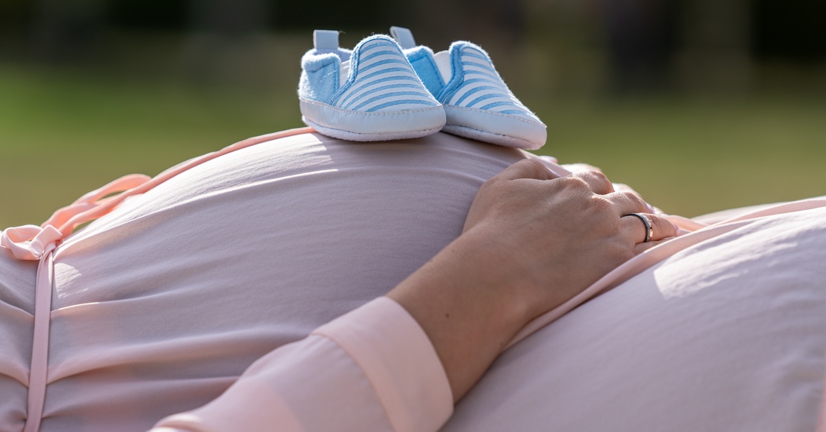 A close-up of a pregnant belly in pink maternity wear with blue-baby shoes on top and hands cradling the bump.