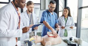 A group of people wearing lab coats and stethoscopes is jotting down notes while examining a training mannequin.