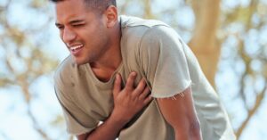 A man wearing a brown shirt and blue pants is clutching his chest in pain while hunched over outdoors.