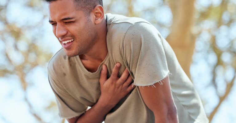 A man wearing a brown shirt and blue pants is clutching his chest in pain while hunched over outdoors.