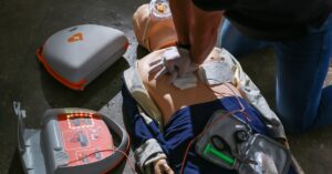 A medical doll lies on the cement as a person performs resuscitation while using an automatic external defibrillator.
