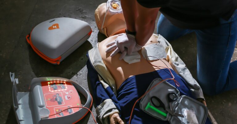 A medical doll lies on the cement as a person performs resuscitation while using an automatic external defibrillator.