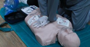 A person performing first aid CPR on a training mannequin while it is attached to an AED device on the floor.