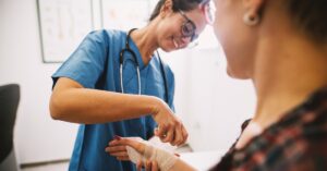 A woman wearing blue scrubs and a stethoscope applies a medical bandage around the hand of another person.