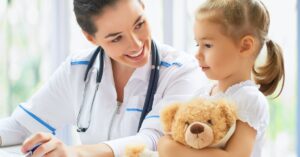 A woman wearing a white lab coat with blue details sits next to a child holding a brown teddy bear. The woman smiles.