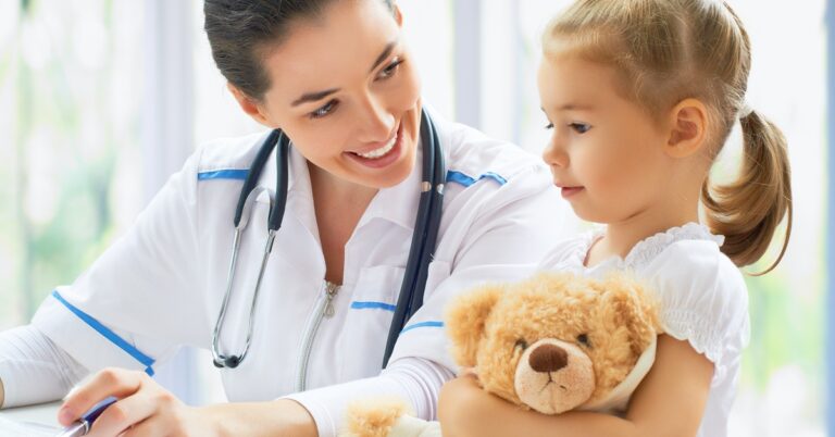 A woman wearing a white lab coat with blue details sits next to a child holding a brown teddy bear. The woman smiles.