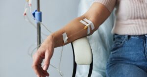A person wearing a pink shirt and blue jeans sits on a white chair with an IV drip treatment on their arm.