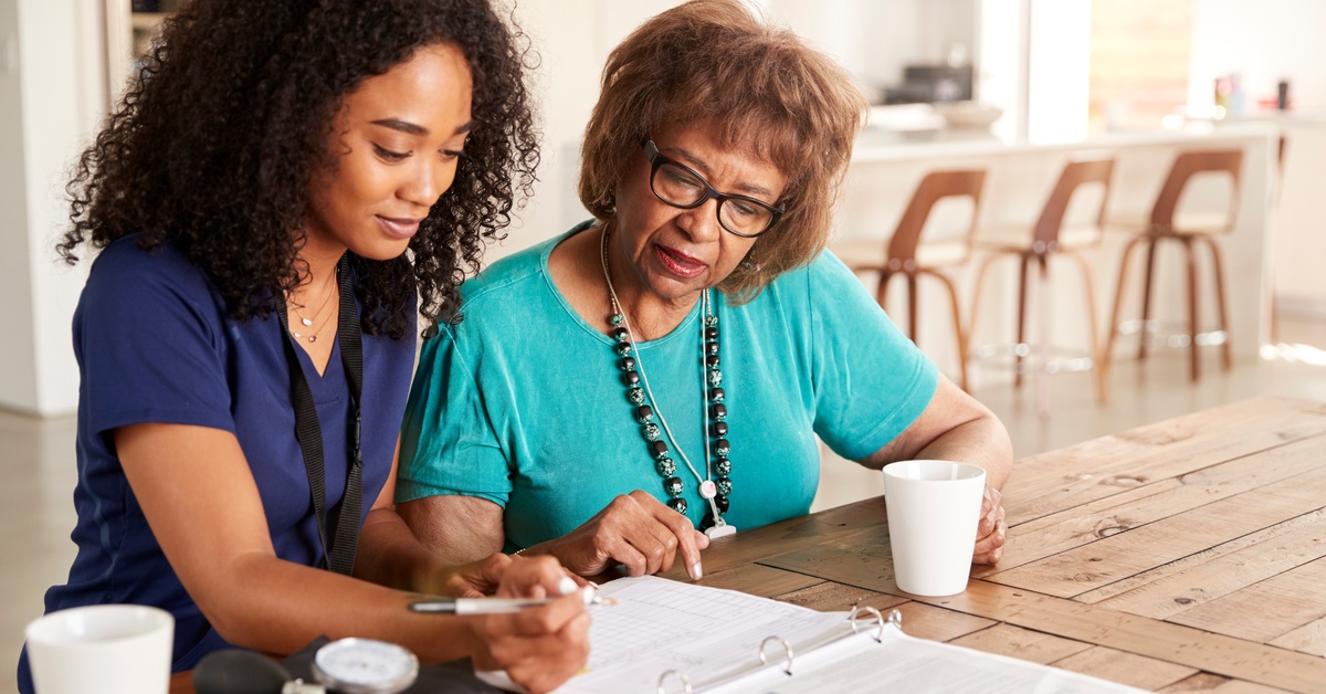 A woman wearing navy blue scrubs sits next to an older woman wearing teal scrubs as they write documents on the table.