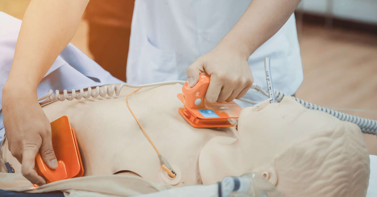 A healthcare professional wearing white scrubs presses two defibrillator electrodes on the chest of a training doll.