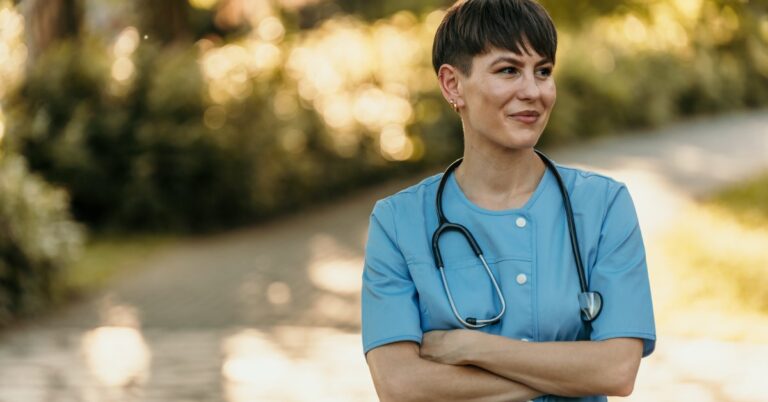 A confident healthcare professional woman nurse standing outdoors in her scrubs, arms crossed and softly smiling.