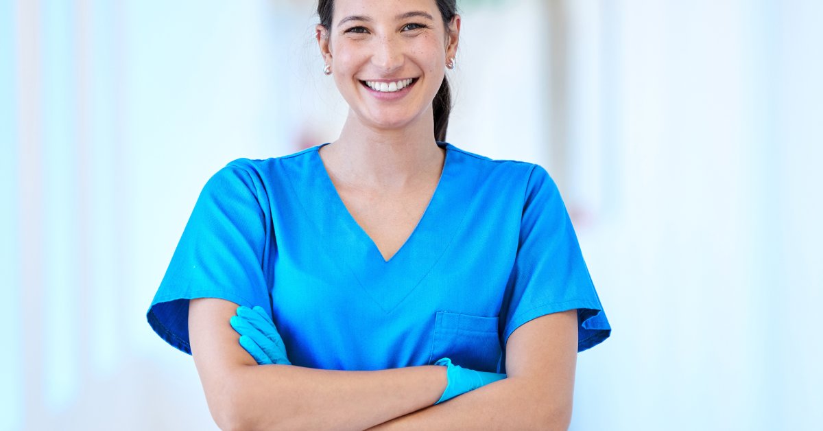A smiling trauma nurse in blue scrubs crosses her arms and stands confidently in a bright hospital corridor.