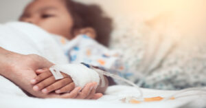 A small child lies in a hospital bed with an IV running through its arm. An adult tenderly holds the child's hand.