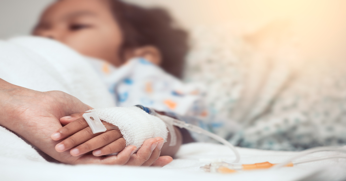 A small child lies in a hospital bed with an IV running through its arm. An adult tenderly holds the child's hand.