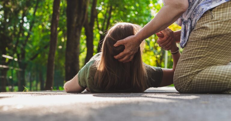 A senior woman on her knees holding the head and the right hand of a woman lying in a concrete path outdoors.