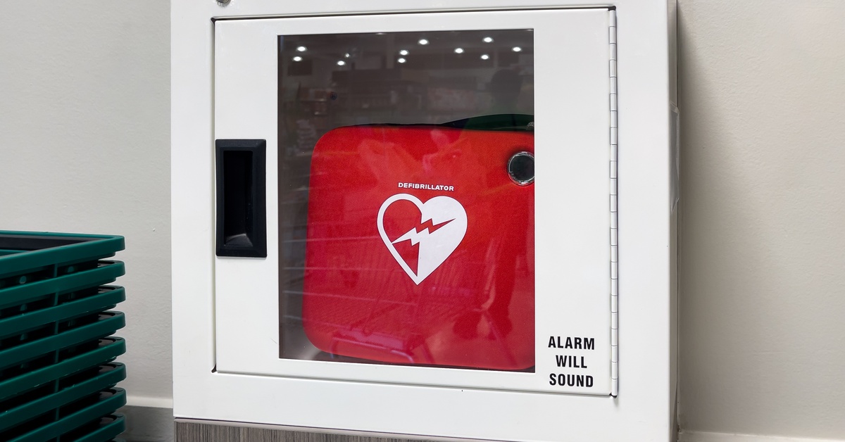 A defibrillator inside a glass case with a black handle, placed on a wall displaying the words “alarm will sound”.