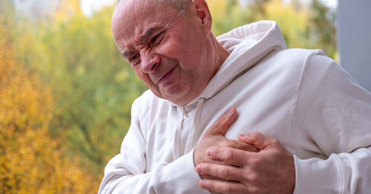 A man wearing a white sweatshirt outdoors, holding both hands on his chest while making a frowny face.