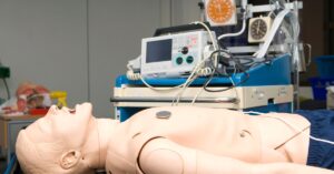 An ACLS training mannequin with a defibrillator and medical equipment in a realistic healthcare environment.