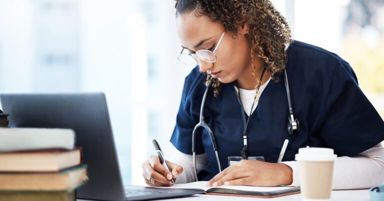A focused nursing school student in scrubs takes notes while studying at a desk with a laptop, books, and coffee.