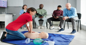 A medical instructor demonstrates CPR techniques on a test dummy as students sit in chairs, intently watching.