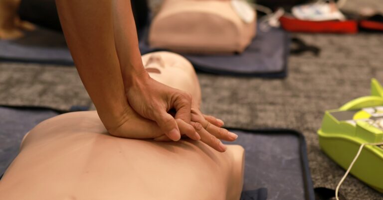 Hands performing chest compressions on a CPR training manikin during a class, with AED training devices visible in the background.