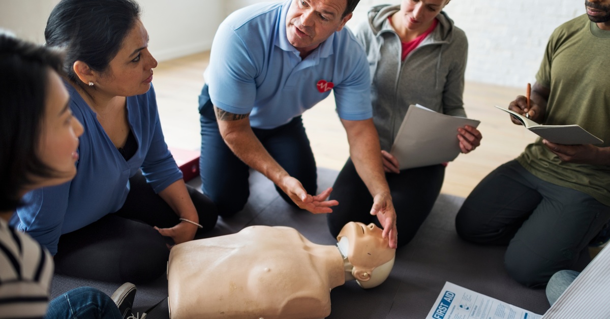 CPR instructor demonstrating airway and assessment techniques on a training manikin while students observe and take notes.