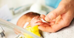 A newborn in a hospital incubator holding an adult caregiver’s finger, showing neonatal care, connection, and support.