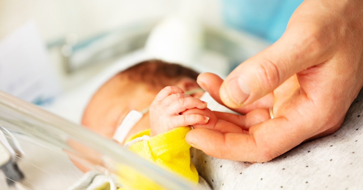 A newborn in a hospital incubator holding an adult caregiver’s finger, showing neonatal care, connection, and support.