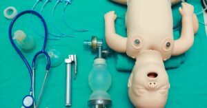 An infant ACLS training mannequin with airway tools, a bag valve mask, a laryngoscope, and a stethoscope on a teal surface.