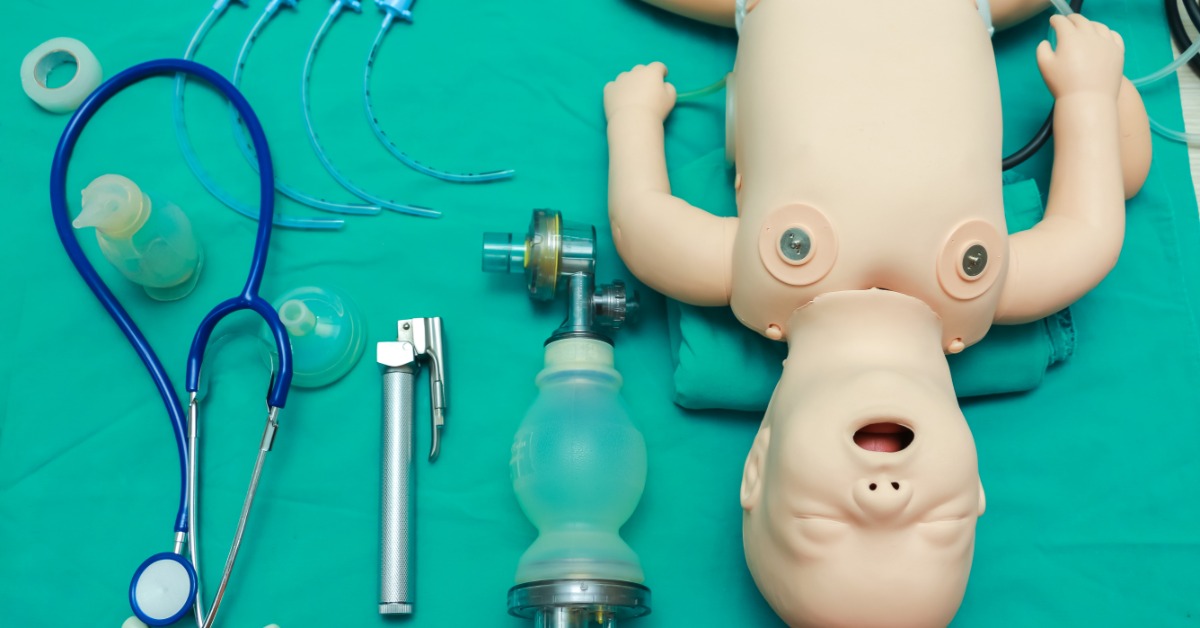 An infant ACLS training mannequin with airway tools, a bag valve mask, a laryngoscope, and a stethoscope on a teal surface.