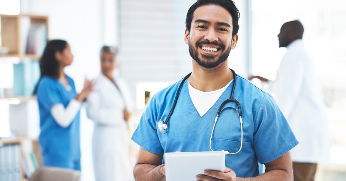 A smiling healthcare provider in blue scrubs holds a tablet as his colleagues engage in a discussion behind him.