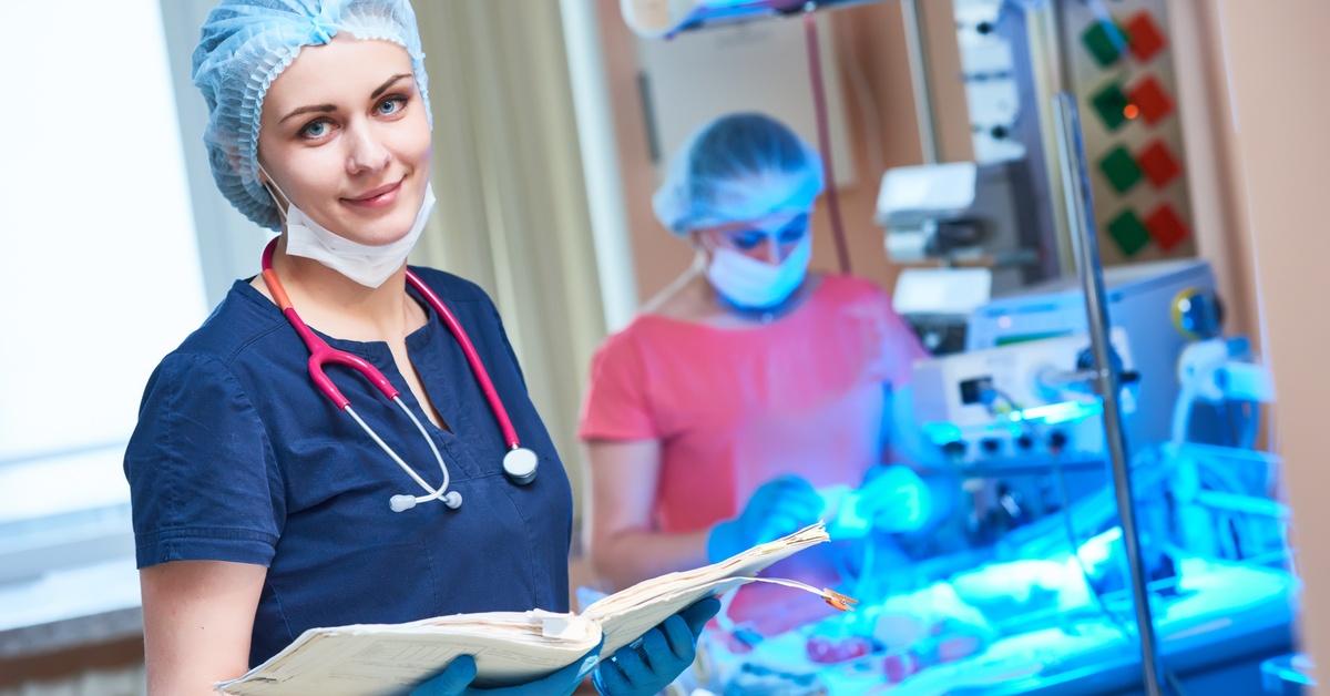 A nurse reviews a patient's chart in the NICU while a colleague provides care to a newborn under incubator lights.