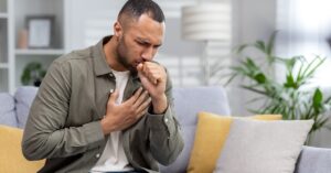 A man sits on a couch, coughing into his fist while holding his chest, displaying signs of respiratory distress at home.